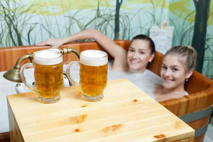 Two smiling women relaxing in a beer bath at Bjórböðin Beer Spa in Iceland, with two frothy beer mugs on a wooden table in the foreground.