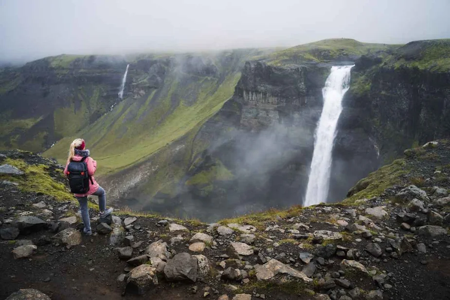 A hiker in a pink jacket and gray pants stands on rocky terrain, gazing at the magnificent Haifoss waterfall in Iceland. The waterfall, one of the highest in the country, cascades down a steep cliff into a misty gorge below. The hiker, wearing a backpack, adds a sense of scale to the scene, highlighting the immense height and power of the falls. The surrounding landscape is lush and green, with patches of fog and low-hanging clouds adding to the mystical atmosphere. In the distance, another smaller waterfall can be seen amidst the rugged cliffs. The image captures the raw beauty and majesty of Iceland's natural wonders.