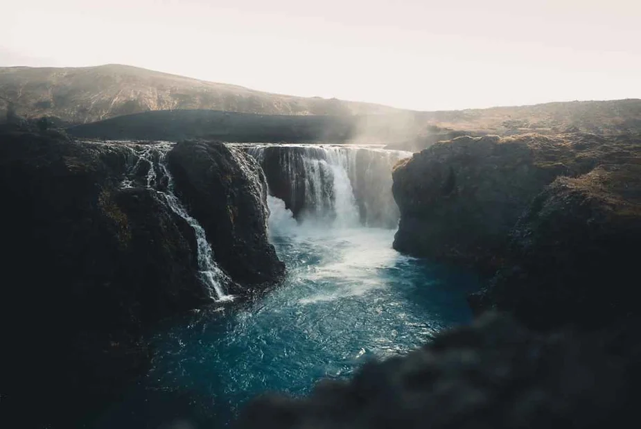 A stunning waterfall cascading into a deep, blue pool surrounded by dark rocky cliffs in the Icelandic highlands. The water, falling gracefully over the rocks, creates a serene and mystical atmosphere, enhanced by the soft light of dawn or dusk. The mist rising from the falls blends with the light, adding a sense of ethereal beauty to the scene. The background reveals rugged terrain, typical of Iceland's dramatic landscapes, with hills and mountains faintly visible in the distance. The overall mood of the image is calm and awe-inspiring, showcasing the natural beauty and tranquility of this remote location.