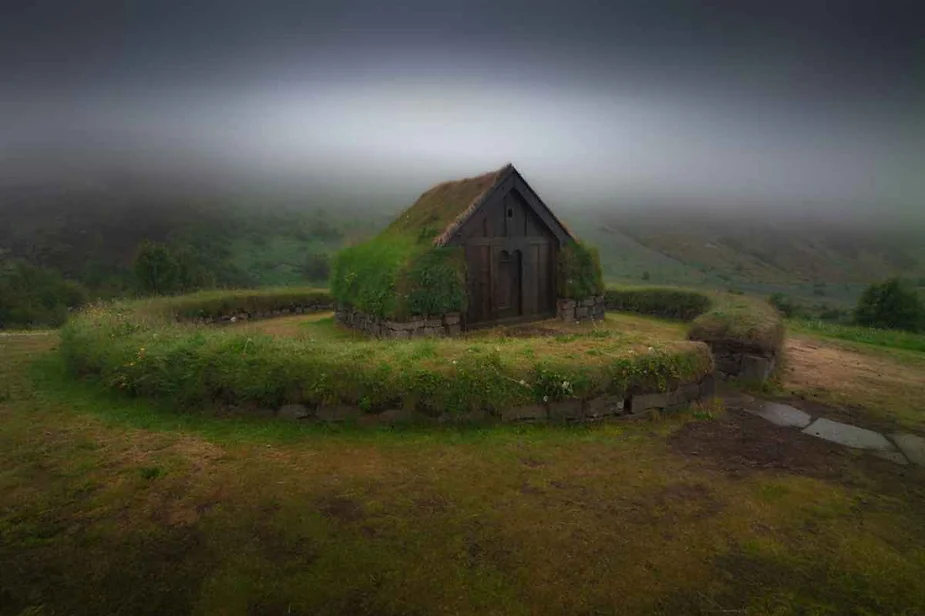 A traditional Icelandic turf house stands solitary in a misty landscape. The small, quaint structure is built with thick walls covered in grass and moss, blending harmoniously with the surrounding greenery. The roof is also made of turf, giving the house a natural, earthy appearance. The mist in the background creates an ethereal, almost otherworldly atmosphere, obscuring the distant landscape and enhancing the sense of isolation and serenity. The setting is peaceful, with a soft, muted light filtering through the fog, highlighting the unique architecture and the lush vegetation. This scene captures the essence of Iceland's historical architecture and its integration with nature.