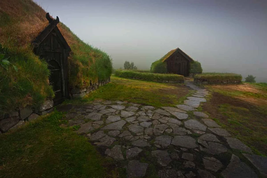 A tranquil scene showcasing traditional Icelandic turf houses enveloped in a thick fog. The houses, with their roofs covered in lush green grass, blend seamlessly with the surrounding landscape. Stone pathways lead to the wooden doors of these quaint structures, enhancing the rustic charm. The fog creates a mystical atmosphere, softening the details and adding an ethereal quality to the scene. The foreground features a well-maintained stone path and vibrant green grass, while the background fades into the mist, suggesting a serene and secluded setting typical of Iceland's unique and historical architecture.