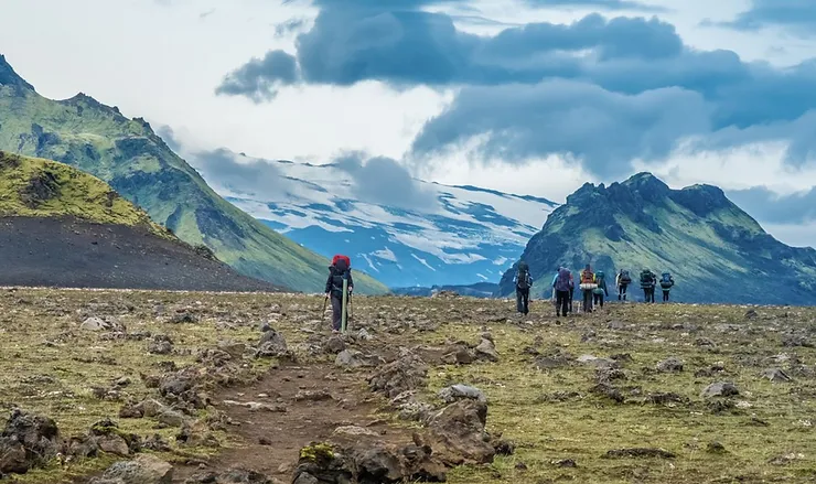 Group of hikers trekking through a rugged, mountainous landscape in Iceland, with snow-capped peaks and dramatic clouds in the background.