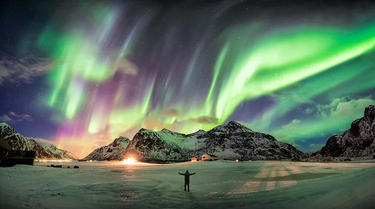 A person standing with arms outstretched, gazing at the vibrant Northern Lights dancing across the night sky over a snowy landscape with mountains in the background.