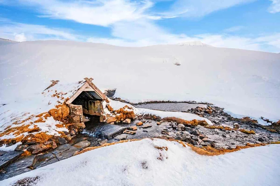 A small hot spring nestled in the Icelandic landscape, partially covered in snow. The spring is surrounded by rocks and features a small, rustic shelter built with wooden beams and stone walls, blending naturally into the snowy environment. The warm water from the spring creates steam that contrasts with the cold, snowy surroundings. The area is peaceful, with snow-covered hills and a clear blue sky above, creating a serene and picturesque winter scene. This image captures the unique beauty of Icelandic hot springs, especially during the winter season, highlighting the contrast between the warmth of the geothermal waters and the cold, snowy landscape.