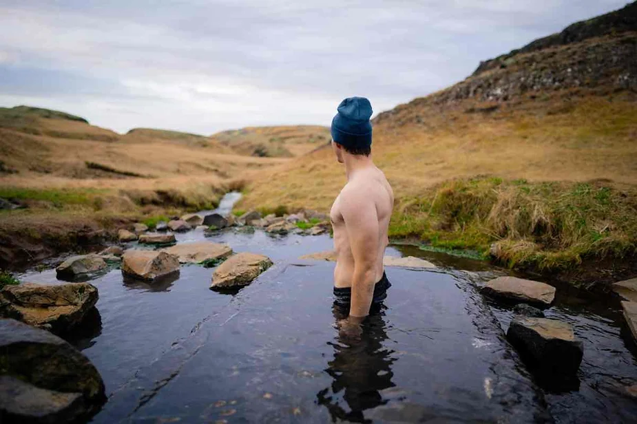 A man with a beanie is seen from behind, standing waist-deep in a natural hot spring surrounded by rocks in the Icelandic countryside. The serene landscape, characterized by rolling hills covered in golden grass, extends into the background. The man is immersed in the warm water, enjoying the peaceful and natural setting, which offers a perfect retreat for relaxation. The sky is overcast, adding to the tranquil and introspective atmosphere of the scene. This image captures the unique experience of enjoying geothermal waters in a picturesque and remote Icelandic setting.