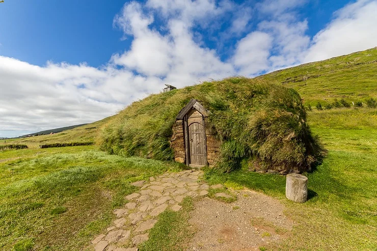 A traditional turf house at Eiriksstadir, Iceland, showcasing its rustic wooden door and roof covered with green grass, set against a backdrop of rolling hills and a blue sky with fluffy clouds.