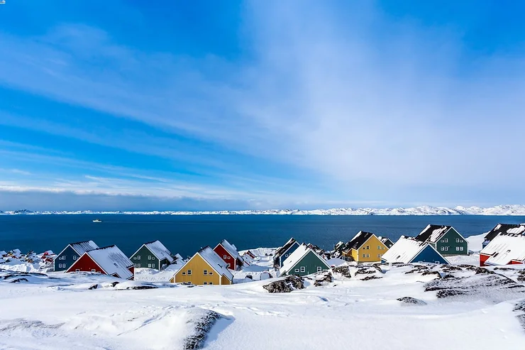 A picturesque view of colorful houses nestled in a snowy landscape by the sea, with a clear blue sky and snow-covered mountains in the distance.