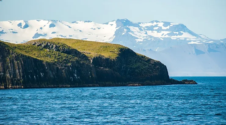 Oxney Island with its rugged coastline and green grass, set against the backdrop of snow-capped mountains under a clear blue sky.