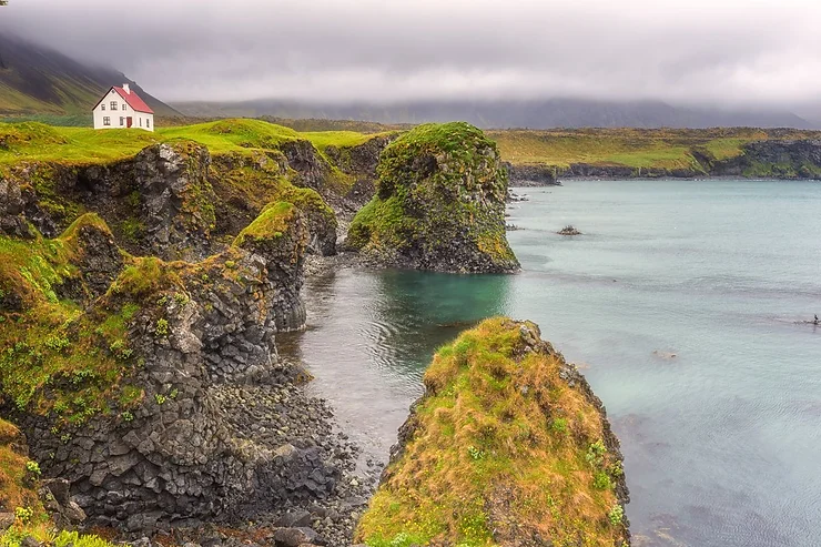 A charming house perched on the green cliffs of Snæfellsnes Peninsula, overlooking the tranquil turquoise waters, with a backdrop of misty mountains.