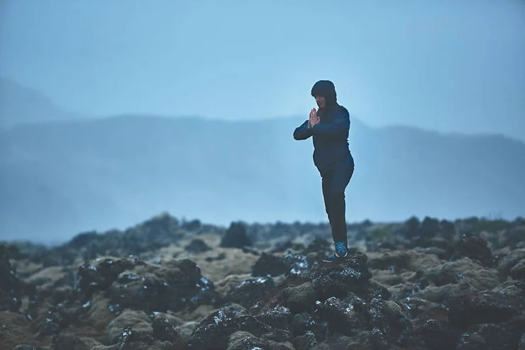 A person practicing yoga in a meditative pose, standing on a rock amidst a rugged and misty Icelandic landscape under a moody, overcast sky.