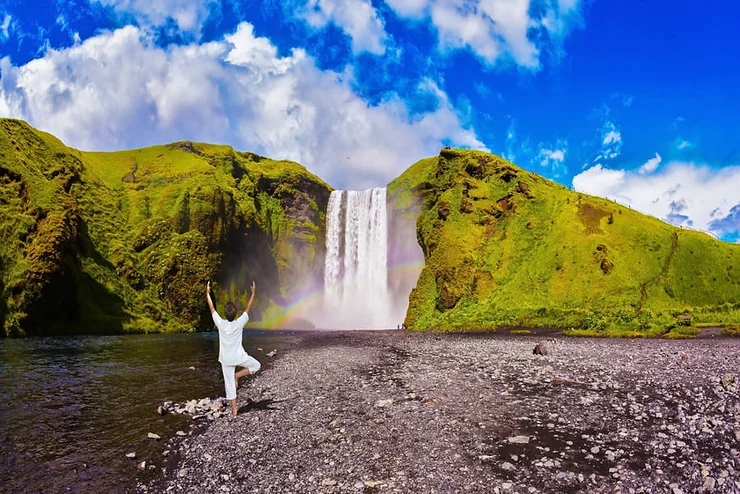 A person practicing yoga in a tree pose near a stunning waterfall in Iceland, surrounded by vibrant green hills and under a bright blue sky with fluffy clouds.