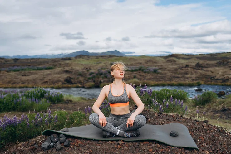 A woman practicing yoga in a serene Icelandic landscape, sitting cross-legged on a yoga mat near a river, surrounded by purple wildflowers and rugged terrain under a cloudy sky.