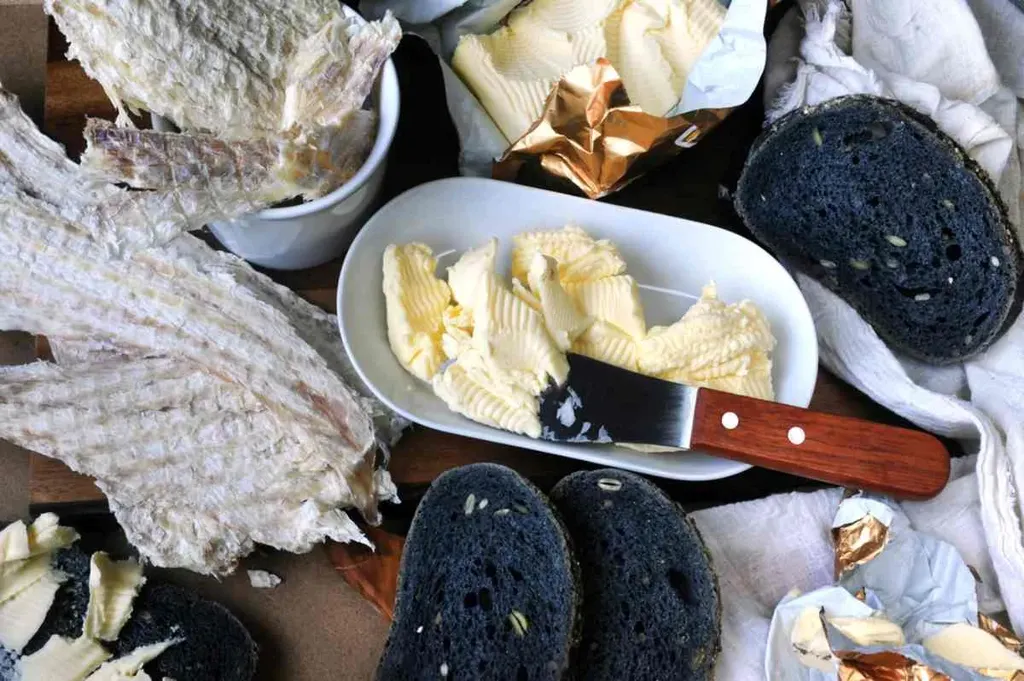 A traditional Icelandic dish featuring hardfiskur (dried fish), accompanied by butter and dark bread. The hardfiskur, a popular Icelandic snack, is served in flaky, dried pieces, showcasing its light, slightly translucent texture. A dish of creamy butter with a knife ready for spreading adds a rich, smooth element to the meal. The dark bread, a distinctive Icelandic rye bread known as rúgbrauð, is cut into slices and pairs perfectly with the fish and butter, offering a hearty and unique culinary experience. The combination highlights Iceland's traditional flavors and simple yet satisfying cuisine.