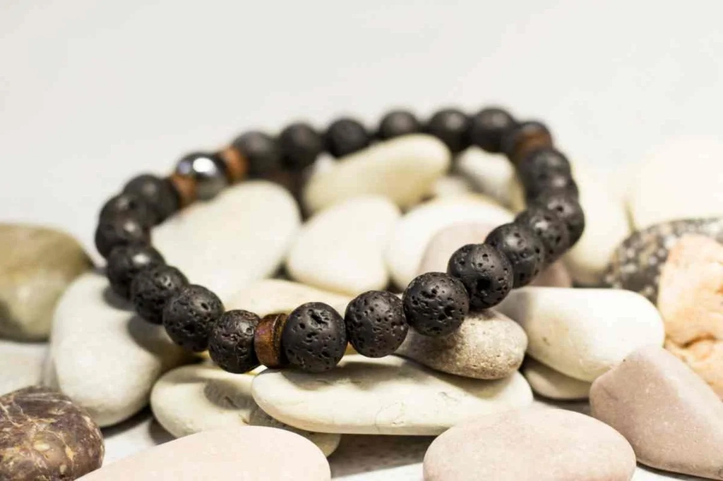 A close-up of a stylish lava bead bracelet placed on a bed of smooth, white pebbles. The bracelet is made of round, black lava stones, each with a unique, porous texture that adds an earthy, rustic charm. This type of jewelry is popular in Iceland, often worn as a fashion accessory or used in aromatherapy due to the beads' ability to absorb essential oils. The contrast between the dark lava stones and the light-colored pebbles highlights the bracelet's natural beauty.