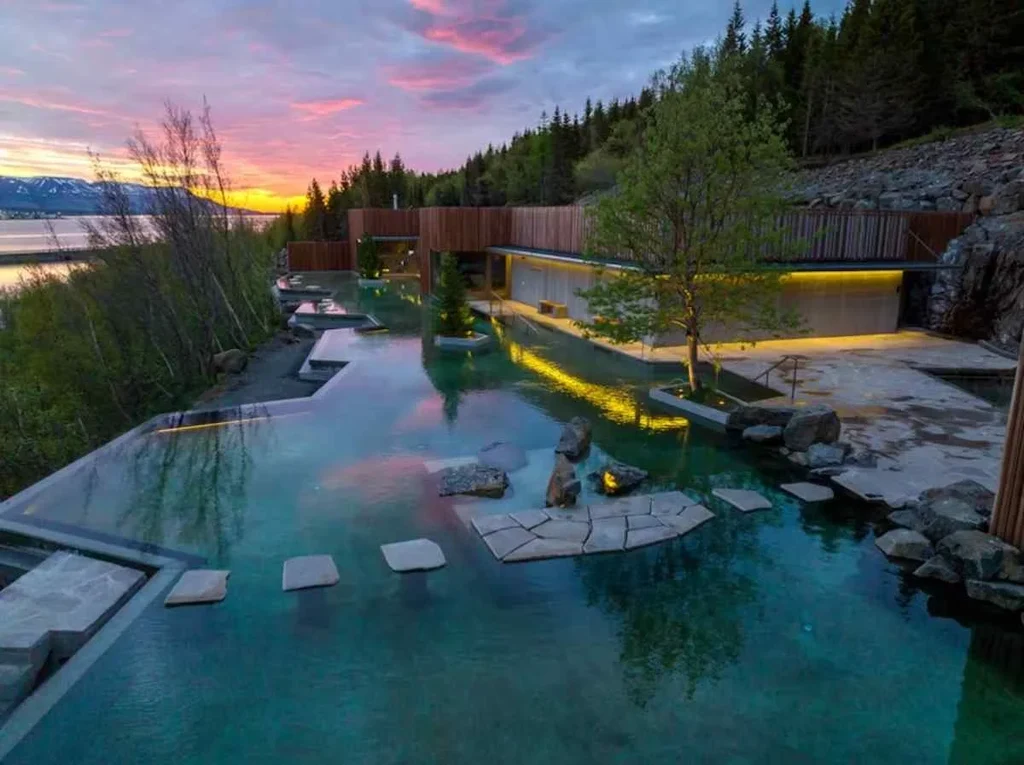 A serene view of the Forest Lagoon in Iceland at sunset. The lagoon's clear, calm waters reflect the surrounding forest and a stunning, colorful sky. The modern facility features multiple pools with natural stone pathways and greenery, blending seamlessly into the environment. Soft lighting from the building adds warmth to the scene, highlighting the minimalistic and tranquil atmosphere. In the background, trees line the lagoon, and a distant mountain range is visible, creating a picturesque and peaceful setting.