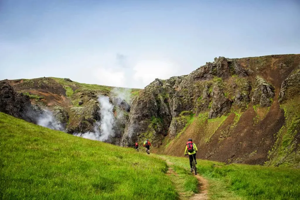 A group of cyclists rides along a narrow dirt path through the lush green countryside of Iceland.