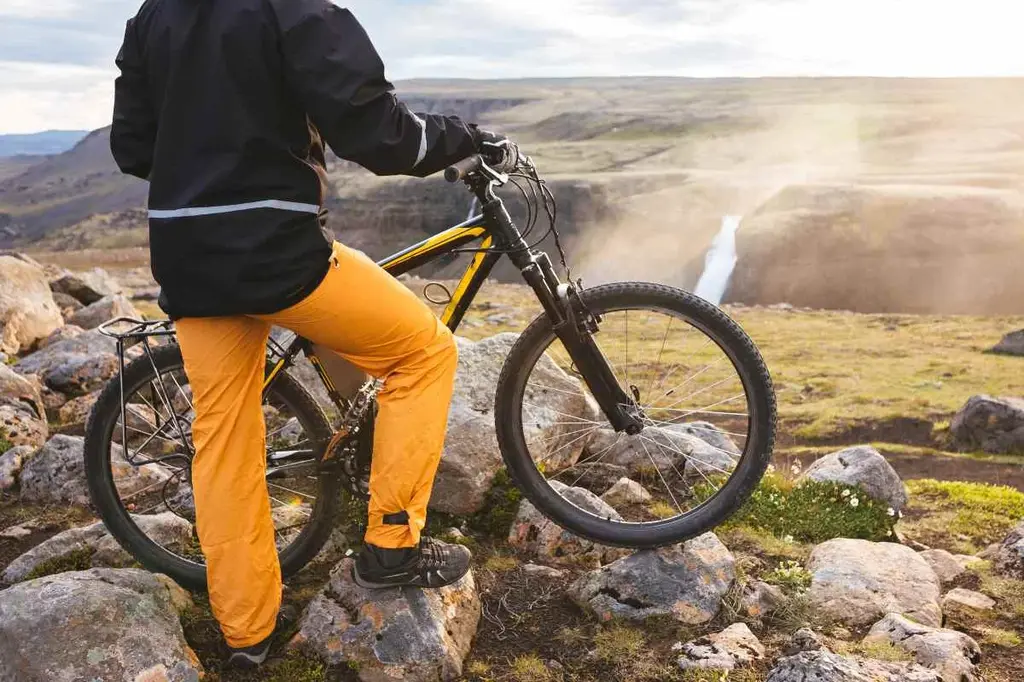 A cyclist in yellow pants and a black jacket stands with their mountain bike on rocky terrain, overlooking a stunning Icelandic landscape.