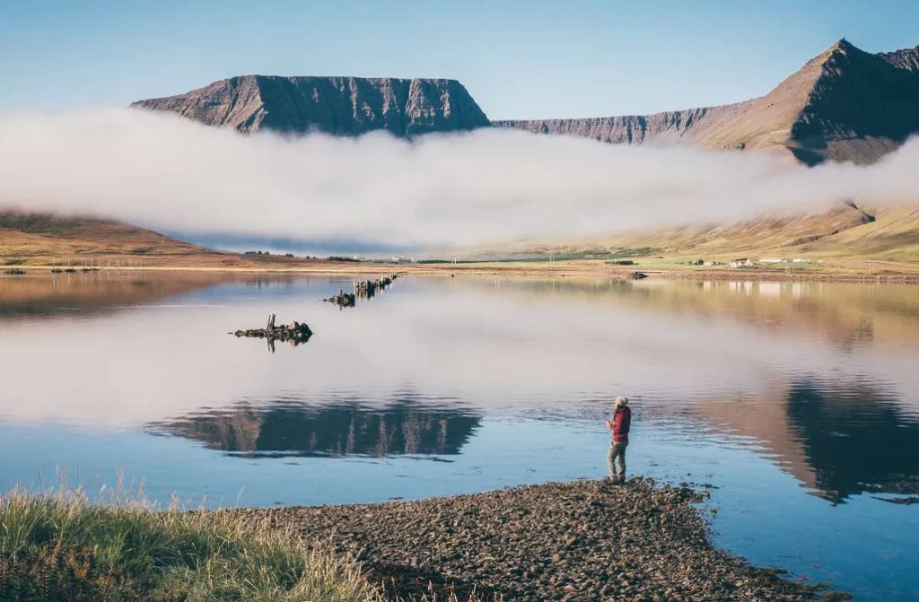 A cyclist stands near the serene waters of a fjord in Iceland's Westfjords region.