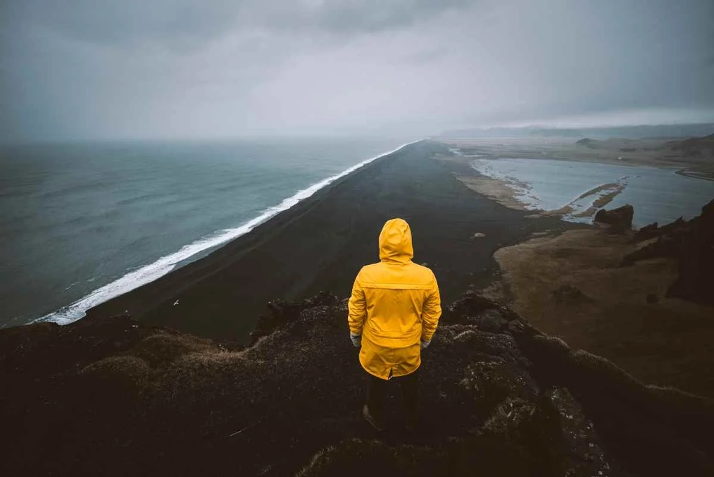 A person in a yellow raincoat stands at the edge of a cliff, gazing out over a dramatic, dark sandy coastline that stretches into the distance. The ocean waves crash against the shore under a cloudy, overcast sky, creating a moody and atmospheric scene. The vast landscape captures the rugged beauty of Iceland's natural environment, with the solitary figure embodying a sense of exploration and adventure. The image conveys a moment of quiet reflection and awe at the power and majesty of nature.