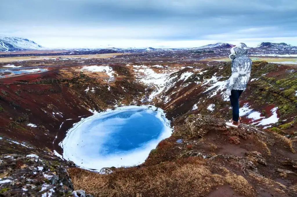 A stunning view of Kerið Crater in Iceland, featuring a visitor in a winter jacket standing at the edge of the crater. The scene captures the vibrant colors of the volcanic landscape, with rich red and brown tones contrasting against the frozen blue lake at the crater's bottom. Snow patches and a vast landscape stretch into the distance, showcasing the rugged and serene beauty of Iceland's Golden Circle. The overcast sky adds a mystical atmosphere to this breathtaking natural wonder.