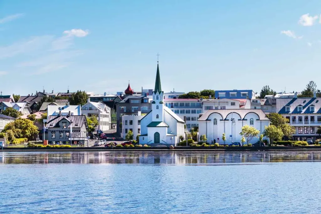 A picturesque view of Reykjavik, Iceland's capital, showcasing its charming waterfront. The scene features a quaint white church with a green spire, surrounded by colorful and modern buildings. The calm blue waters of the lake in the foreground reflect the clear, sunny sky and the vibrant architecture. This image captures the serene and welcoming atmosphere of Reykjavik, highlighting its unique blend of traditional and contemporary styles.