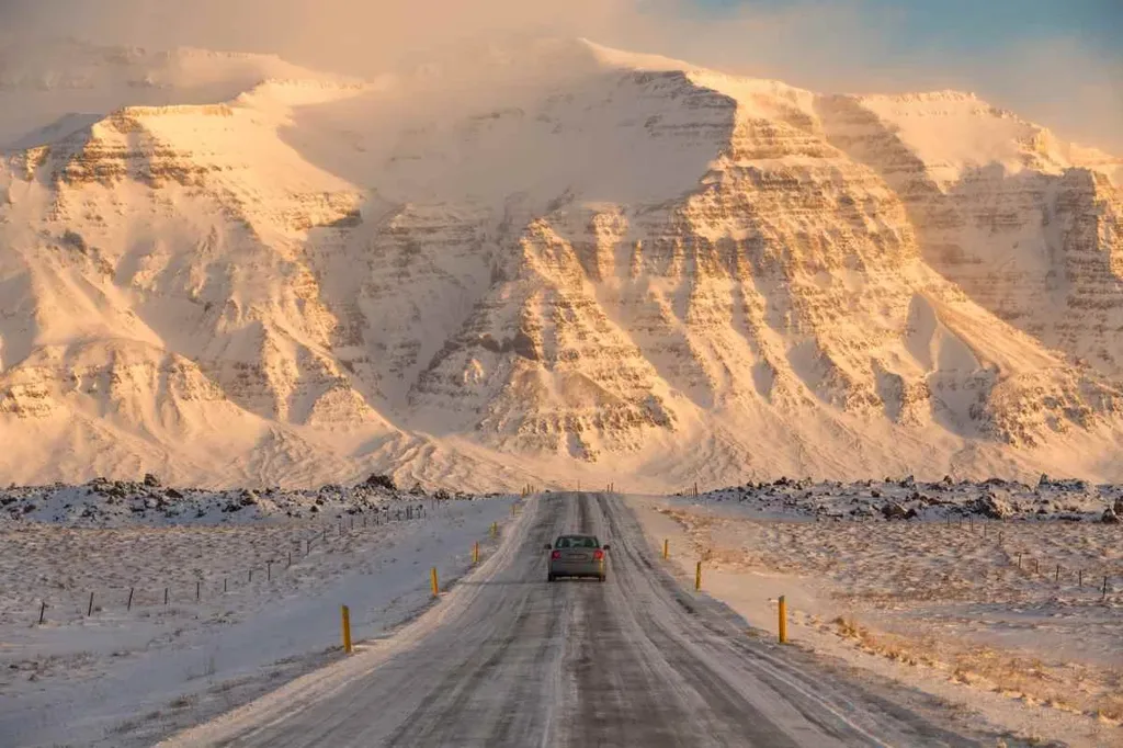 A car drives down a snow-covered road in Iceland, flanked by yellow markers. The road leads toward a dramatic mountain range, bathed in golden light from the setting sun. The towering mountains are blanketed in snow, their rugged features highlighted by the low sunlight. The scene captures the beauty and challenge of driving in Iceland's winter conditions, emphasizing the stark and majestic landscape.