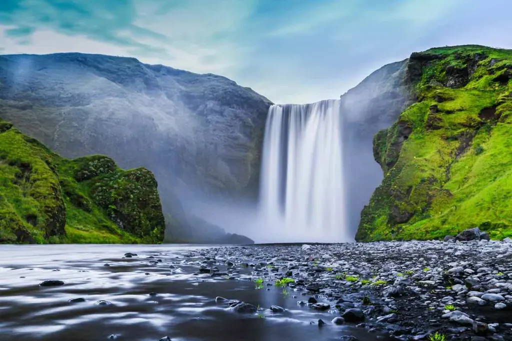 A stunning view of Skogafoss, one of Iceland's most famous waterfalls, cascading down from a high cliff. The powerful waterfall is surrounded by lush green cliffs, with mist rising from the base where the water hits the ground. The foreground features a shallow riverbed with smooth pebbles and vibrant green moss, leading the eye towards the majestic waterfall. The scene is tranquil and awe-inspiring, capturing the natural beauty and serene atmosphere of this iconic Icelandic landscape.