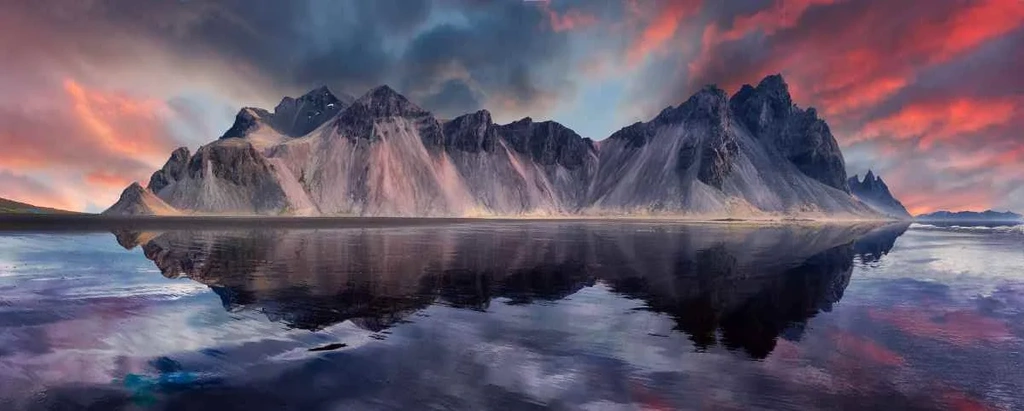 A breathtaking panorama of Stokknes Beach with the dramatic Vestrahorn Mountain in the background. The image captures the serene reflection of the mountain range on the calm waters of the beach, with sharp peaks and ridges cutting through the sky. The sky above is painted with vivid hues of pink, orange, and purple from the setting sun, adding a magical ambiance to the scene. The tranquil water mirrors the vibrant colors and majestic landscape, creating a perfect symmetry and enhancing the sense of tranquility and beauty of this iconic Icelandic location.