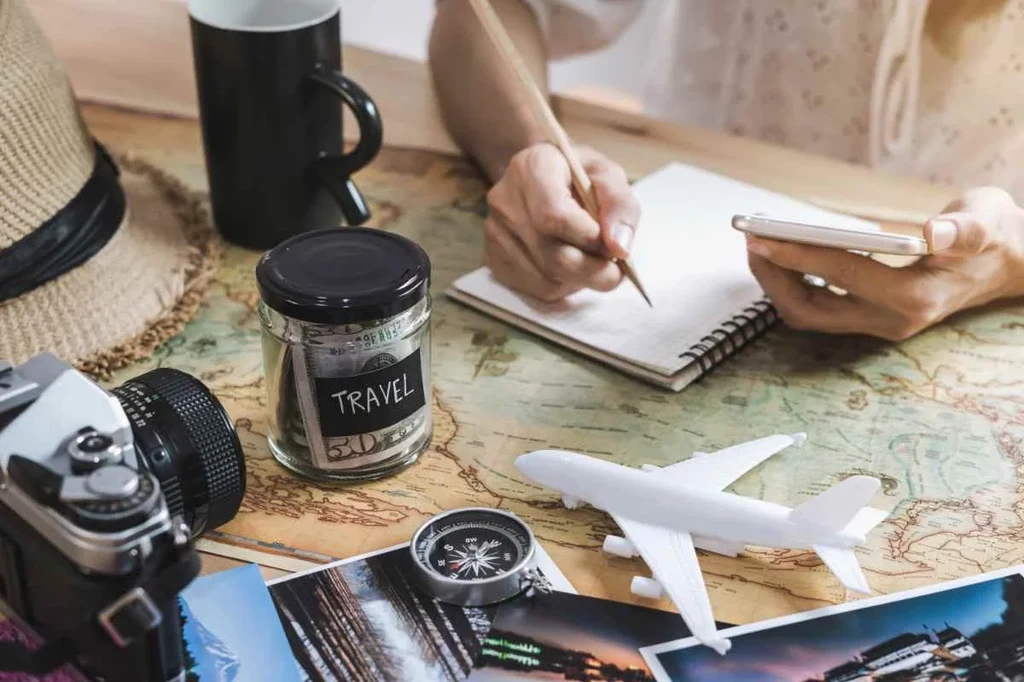 A scene depicting trip planning for Iceland. The photo shows a table with various items, including a glass jar labeled 'travel' filled with money, a vintage camera, small white model airplane, compass, and photographs. A person is seen writing in a notebook with one hand while holding a smartphone in the other. The background features a world map, highlighting the preparation and budgeting for an upcoming journey. The image captures the excitement and careful planning involved in organizing a trip to Iceland.