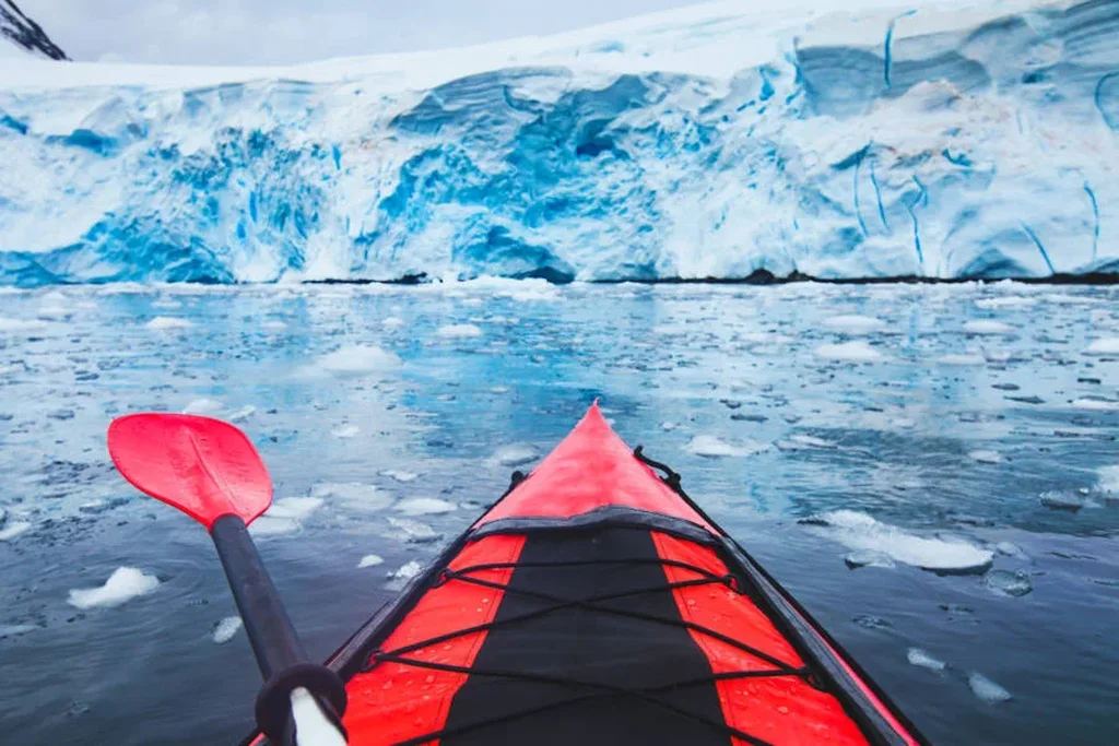 A vibrant red kayak glides through icy waters, surrounded by floating ice chunks. The kayak points towards a massive blue glacier, showcasing its intricate layers and deep crevices. The paddle, matching the kayak's color, is positioned at the side, adding a splash of color to the serene, frozen landscape. The image captures the adventurous spirit of kayaking in Iceland, highlighting the stark beauty and unique challenges of exploring icy waters near glaciers.