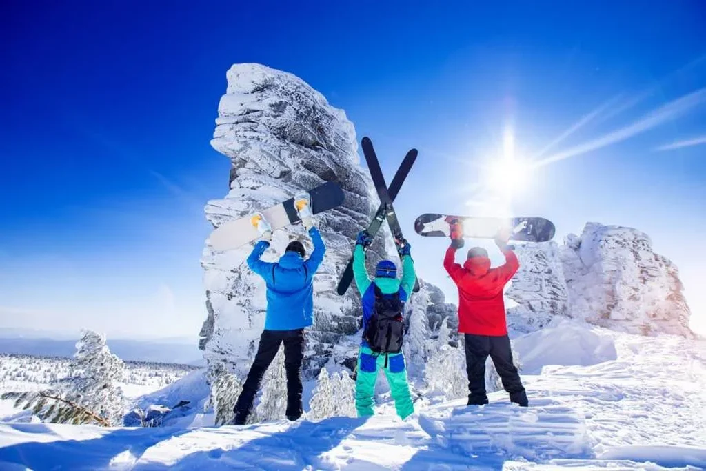 Three winter sports enthusiasts stand triumphantly with their snowboards and skis raised high above their heads against a backdrop of snow-covered mountains in Iceland. They are dressed in bright winter gear, with a clear blue sky and the sun shining brightly. The scene captures the excitement and joy of skiing and snowboarding in Iceland's stunning winter landscape, with snow-covered rocks and trees adding to the pristine beauty of the location.