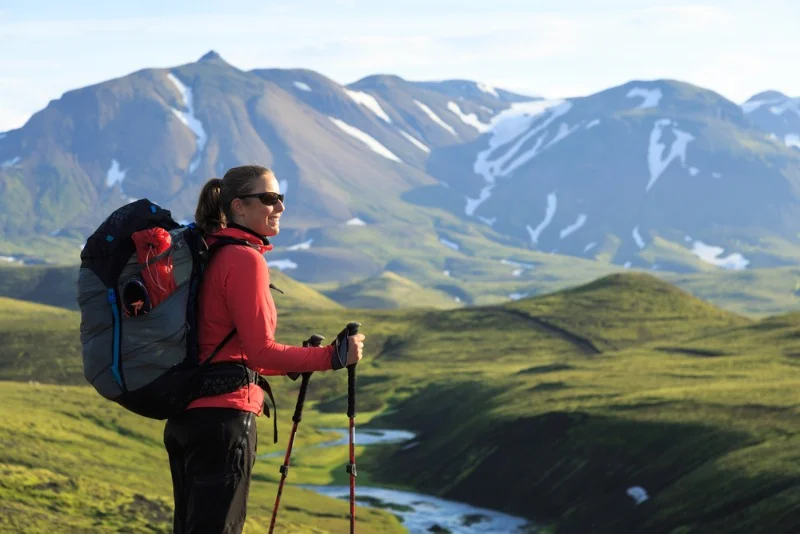 A hiker standing on a green hill, overlooking the stunning landscape of Landmannalaugar, Iceland. The hiker, dressed in a red jacket and black pants, carries a large backpack and uses trekking poles. The background features lush green hills and majestic mountains partially covered with snow. The hiker looks content and enjoys the beautiful natural surroundings, with the bright and clear sky adding to the serene atmosphere. The scene captures the spirit of adventure and the breathtaking scenery of Iceland's highlands, making it an ideal destination for outdoor enthusiasts and hikers.