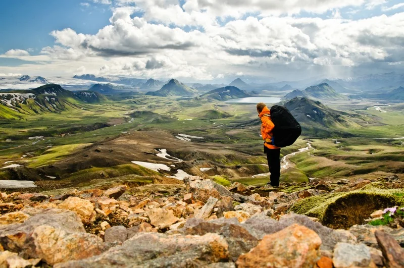 A hiker dressed in an orange jacket and dark pants stands on rocky terrain, overlooking a vast and lush Icelandic landscape. The person carries a large backpack and appears to be taking in the stunning panoramic view. The scene includes rolling green valleys, distant snow-capped peaks, and scattered lakes, all under a partly cloudy sky. The landscape stretches out toward the horizon, showcasing the natural beauty and grandeur of Iceland's wilderness. The image captures a sense of adventure and exploration, highlighting the scale and diversity of the terrain.