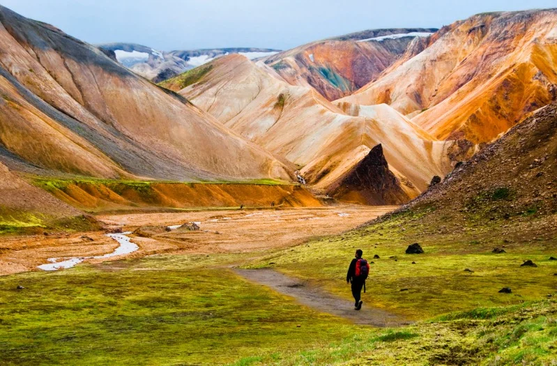 A lone hiker with a red backpack walks along a trail through the vibrant and multicolored hills of Landmannalaugar, Iceland. The landscape features striking orange, red, yellow, and brown rhyolite mountains, creating a stunning and unique natural palette. A small stream winds through the valley, adding to the serene and picturesque scene. The hiker is dressed in dark clothing and stands out against the vivid background, highlighting the scale and beauty of the environment. The image captures the essence of adventure and exploration in one of Iceland's most iconic and colorful landscapes.