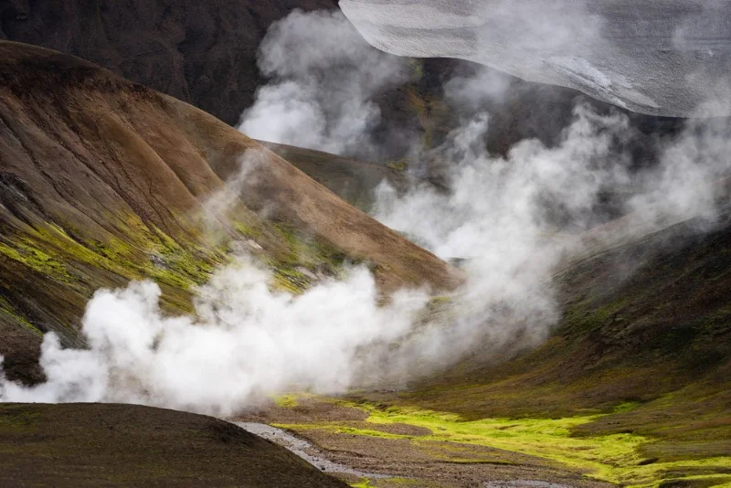 A scenic view of steam vents in Hveradalir, Iceland, showcasing the dramatic geothermal landscape. The image captures a valley with rugged, brown hillsides and vibrant green patches of moss and vegetation. Plumes of steam rise from the ground, creating an ethereal and mystical atmosphere. The geothermal activity is evident with the visible steam and fumaroles dotting the landscape. Above, a rocky outcrop with patches of snow contrasts with the warmer, steam-filled valley below. Hveradalir is known for its geothermal activity, making it a fascinating destination for visitors interested in natural wonders.