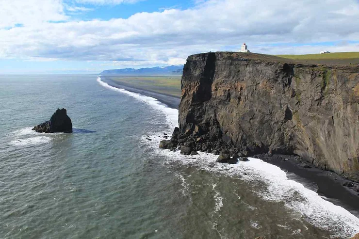 A breathtaking view of the Dyrhólaey Arch in Iceland, with a majestic cliffside overlooking the expansive ocean and a solitary rock formation in the sea, under a partly cloudy sky.