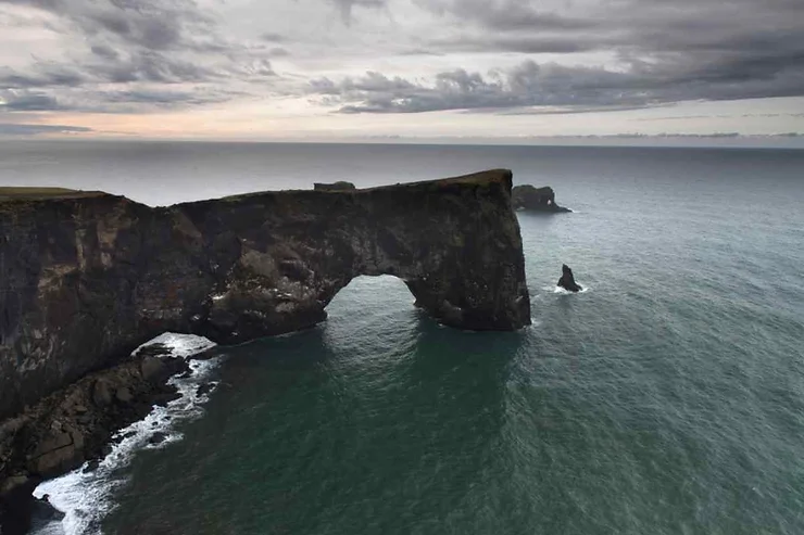 A majestic view of the Dyrhólaey Arch at Dyrhólaey Beach in Iceland, showcasing the natural rock formation jutting into the ocean, with overcast skies and the horizon extending into the distance.