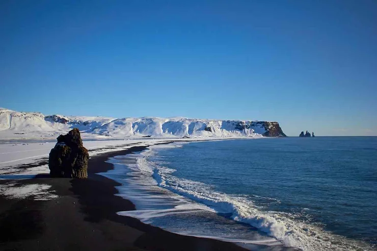 A stunning view of Dyrhólaey Beach in Iceland, featuring black sand contrasting with white snow along the shoreline, with the calm blue ocean extending towards the horizon and snow-covered cliffs in the background under a clear blue sky.