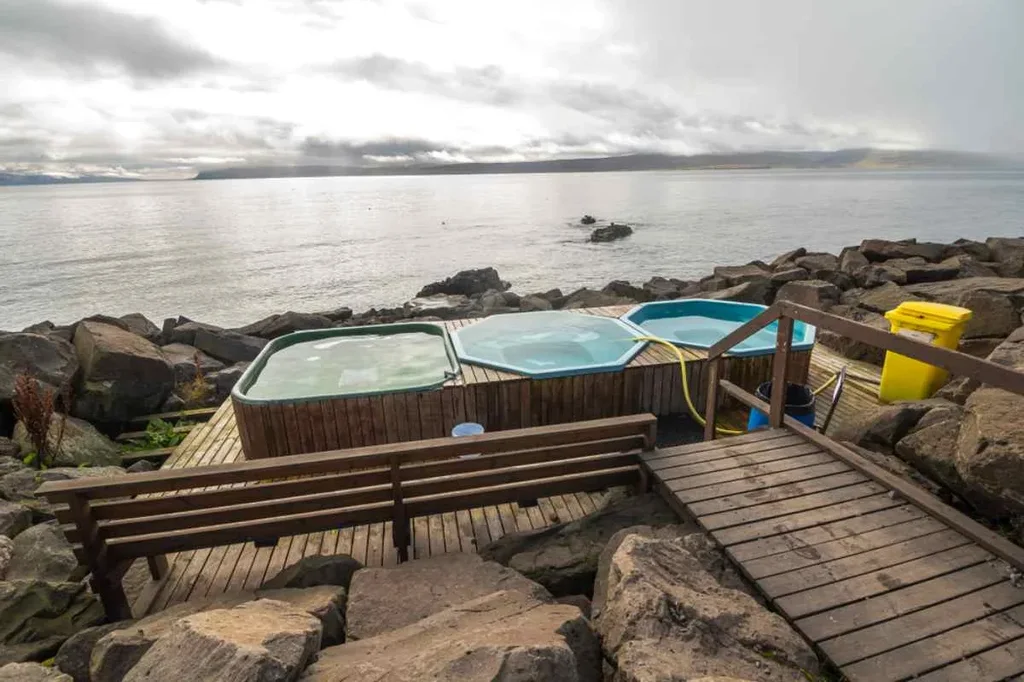 A serene view of the Drangsnes Hot Pots in Iceland, featuring three hot tubs arranged on a wooden platform by the sea.