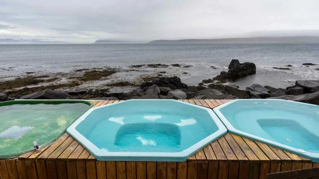 A scenic view of the Drangsnes Hot Pots, featuring three hexagonal hot tubs filled with blue geothermal water. The hot pots are set on a wooden platform by the rocky shoreline, offering a relaxing experience with a backdrop of the calm sea and distant mountains.