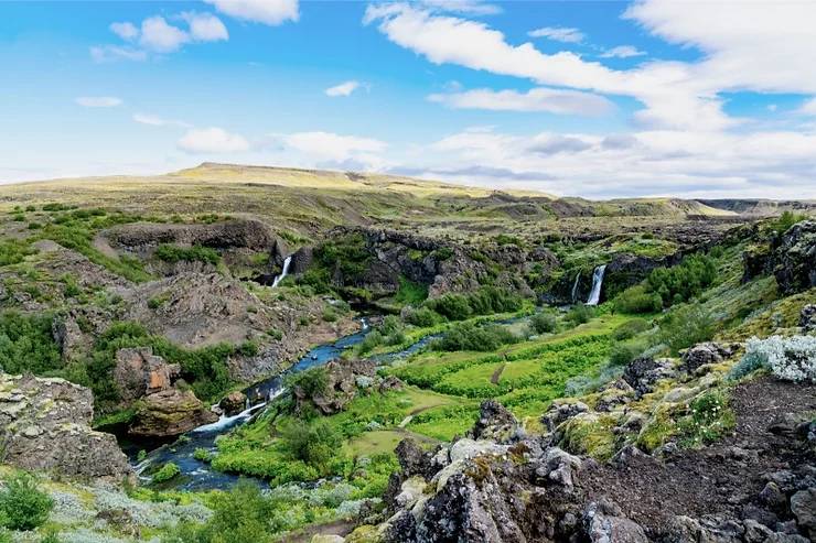 A picturesque view of Gjáin Valley, featuring lush green vegetation, flowing streams, and small waterfalls cascading down rocky cliffs under a bright blue sky with scattered clouds.