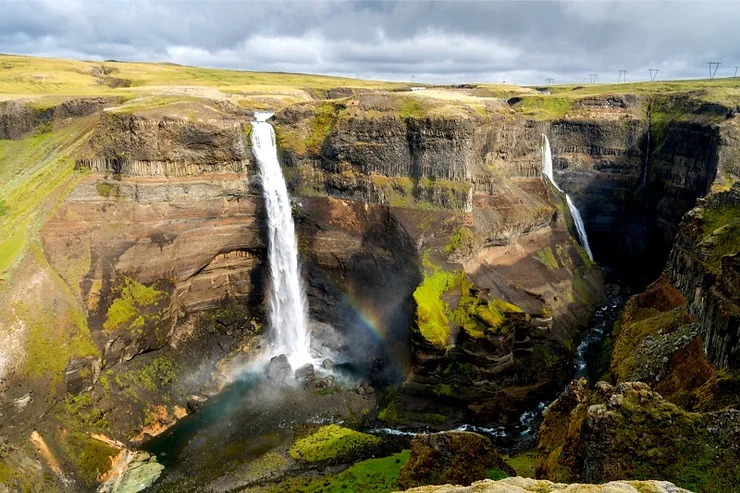 A stunning view of the twin cascades of Háifoss waterfall, plunging dramatically into a deep canyon with a rainbow forming in the mist, surrounded by green and brown rugged cliffs under a cloudy sky.