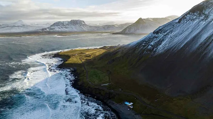 An aerial shot of Krossneslaug, Iceland, featuring dramatic coastal cliffs, turbulent ocean waves, and snowy mountain peaks in the background.