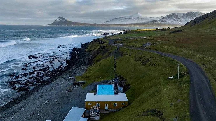 An aerial view of Krossneslaug hot spring nestled on the rugged coastline of Iceland, with waves crashing against the shore and snow-capped mountains in the distance.