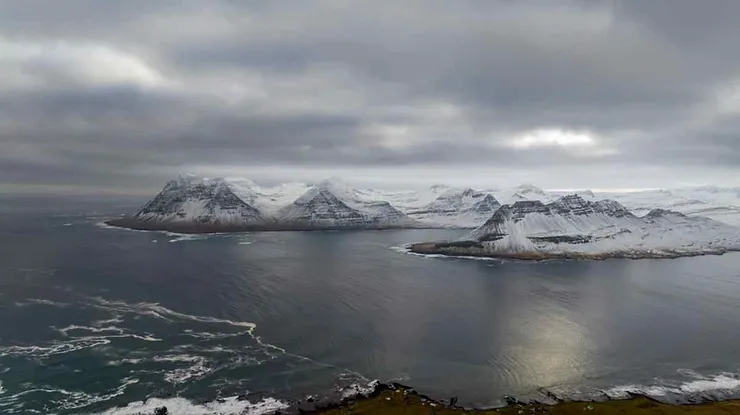 An aerial shot showcasing the rugged coastal mountains near Krossneslaug, Iceland, with snow-capped peaks, overcast skies, and the calm sea reflecting the subdued light.