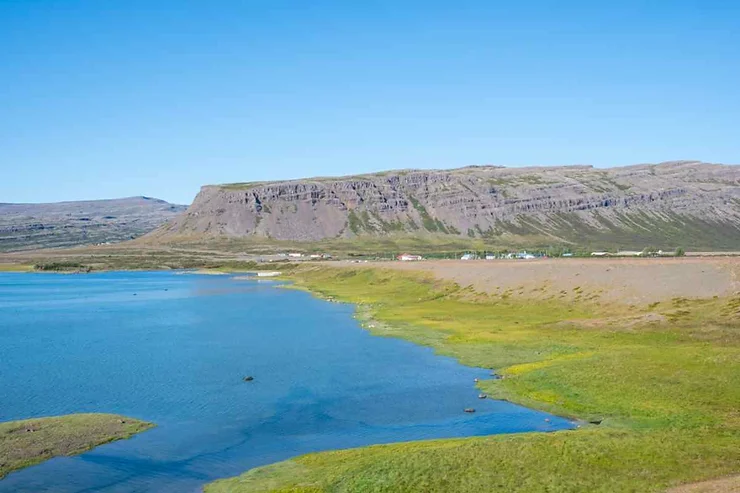 A picturesque landscape showcasing a serene village near Krossneslaug, Iceland, with clear blue waters, lush green fields, and rocky cliffs under a bright blue sky.