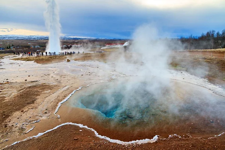 Strokkur Geyser erupts in a powerful jet of water and steam in the Golden Circle, Iceland, with tourists gathered at a safe distance to witness the natural spectacle.