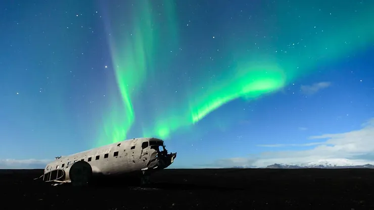 An abandoned plane wreck sits on the black sands of Iceland's South Coast, illuminated by the vibrant green and blue hues of the Northern Lights dancing across the night sky.