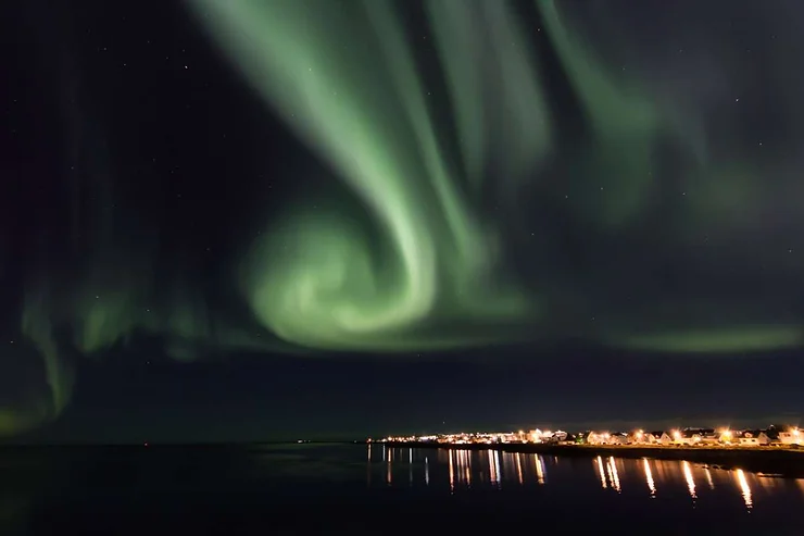 A stunning display of the Northern Lights swirls across the night sky over a small Icelandic town, with reflections of the lights shimmering on the calm water below.