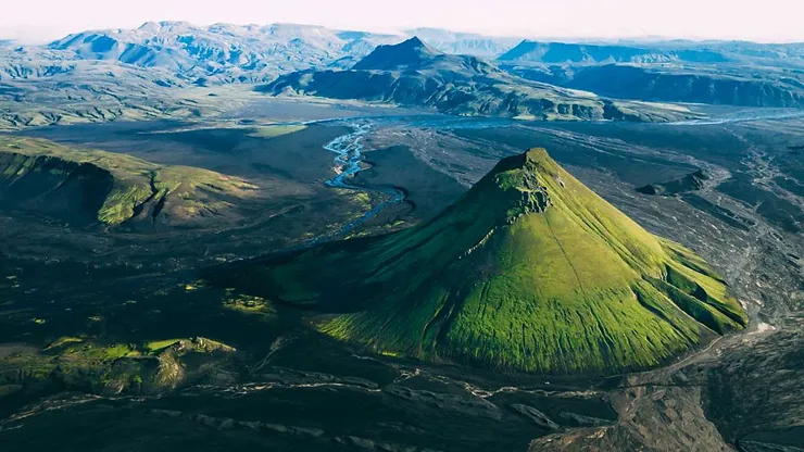 An aerial view of a lush green volcanic cone rising from the surrounding black volcanic landscape, with rugged mountains and winding rivers in the background, showcasing the dramatic terrain of Iceland.